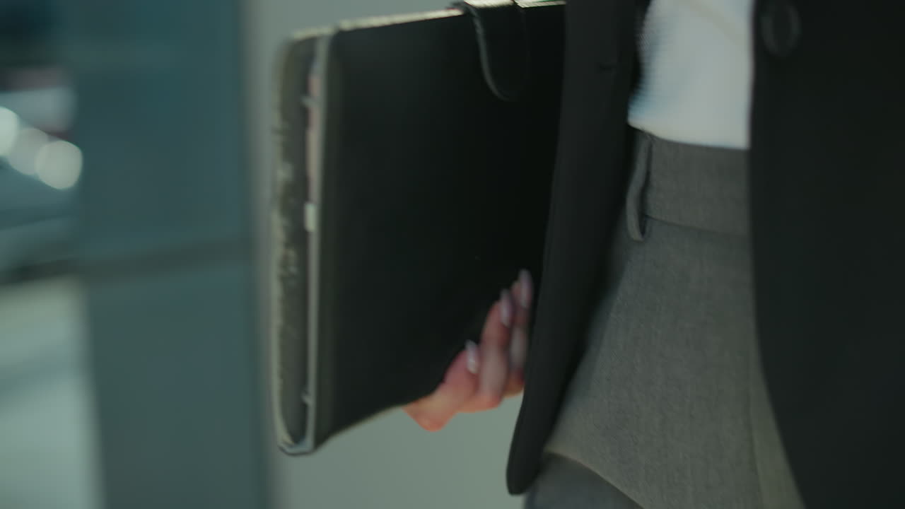 Detailní záběr dámy v černém saku a kalhotách držící tablet, kráčející kolem budovy se skleněnou fasádou odrážející městské rozostřené pozadí v jemném slunečním světle