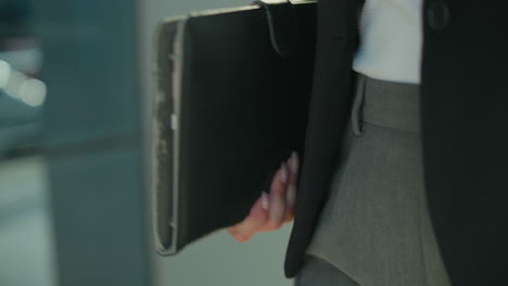 close up waist view of lady in black blazer and trousers holding tab while walking by building with glass facade reflecting urban blur background in soft sunlight