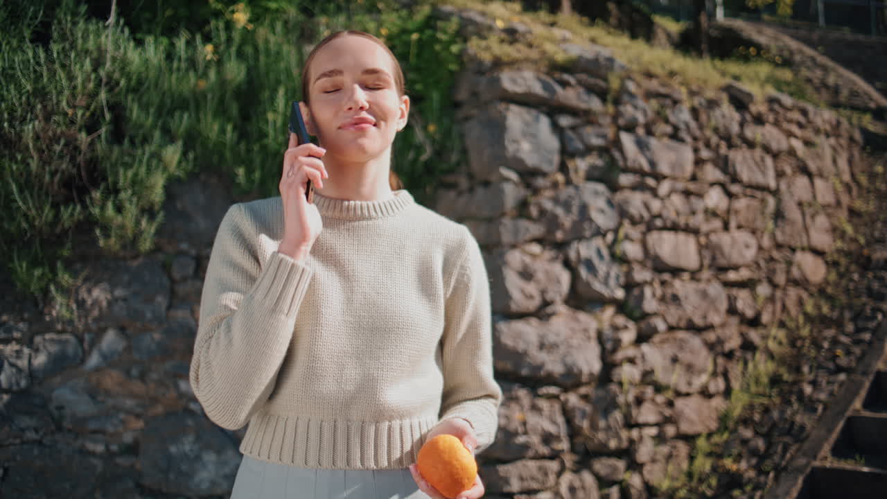 Cute model talking smartphone at greenery park closeup. Woman holding orange