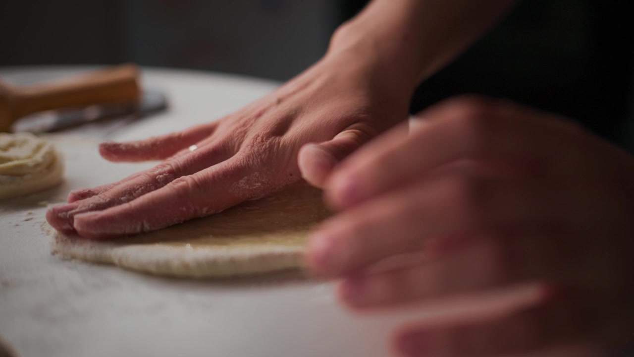 Le mani premono delicatamente l'impasto su una superficie infarinata. L'azione concentrata mostra una preparazione esperta. L'illuminazione calda evidenzia l'impasto testurizzato e lo spazio di lavoro. Ideale per contenuti culinari, di ristorazione o tutorial di cucina.