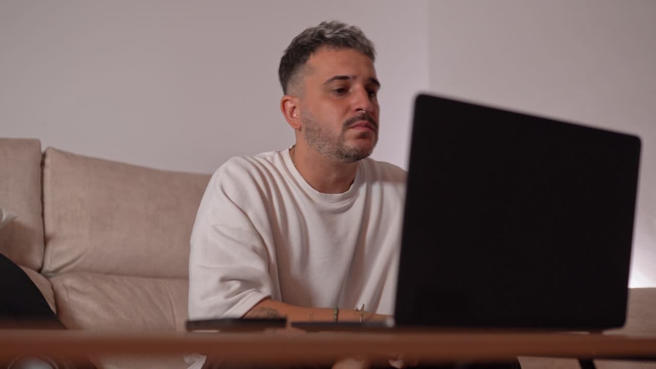 Man sitting on sofa showing signs of frustration with hands on face while working on laptop in modern living room environment