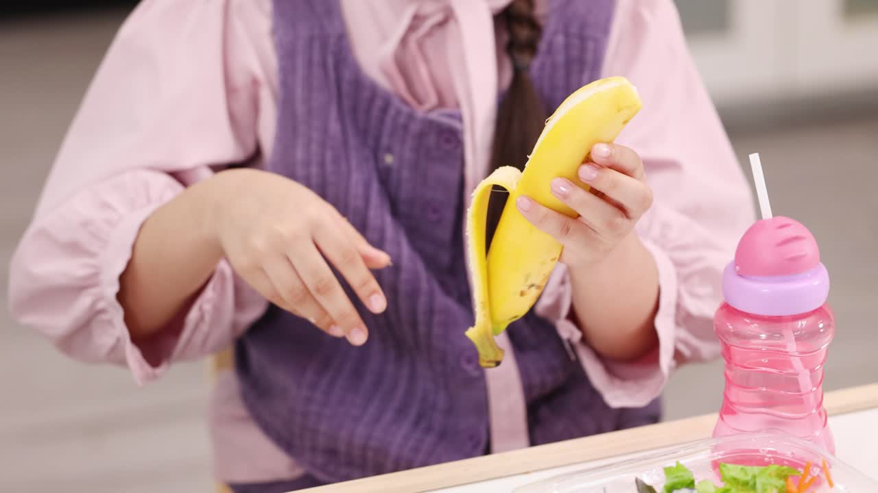 Schoolchild in uniform peels banana, about to eat, under soft indoor lighting, close-up view