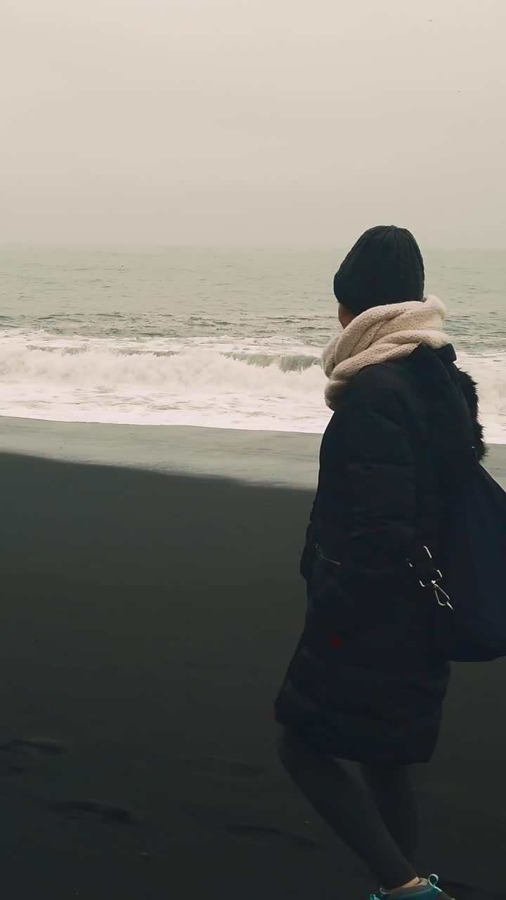Spiaggia di Reynisfjara: donna solitaria in abiti invernali che cammina lungo la costa di una spiaggia di sabbia nera in una giornata nebbiosa. Islanda