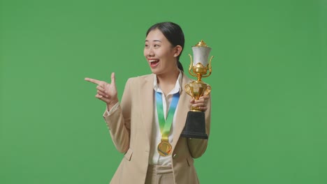 asian business woman in a suit with a gold medal holding a gold trophy in her hands, smiling, and pointing to side on green screen background in the studio