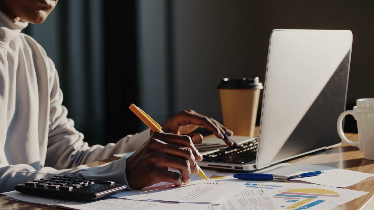 A side-angle video shot of a desk with a laptop, papers, calculator, and coffee