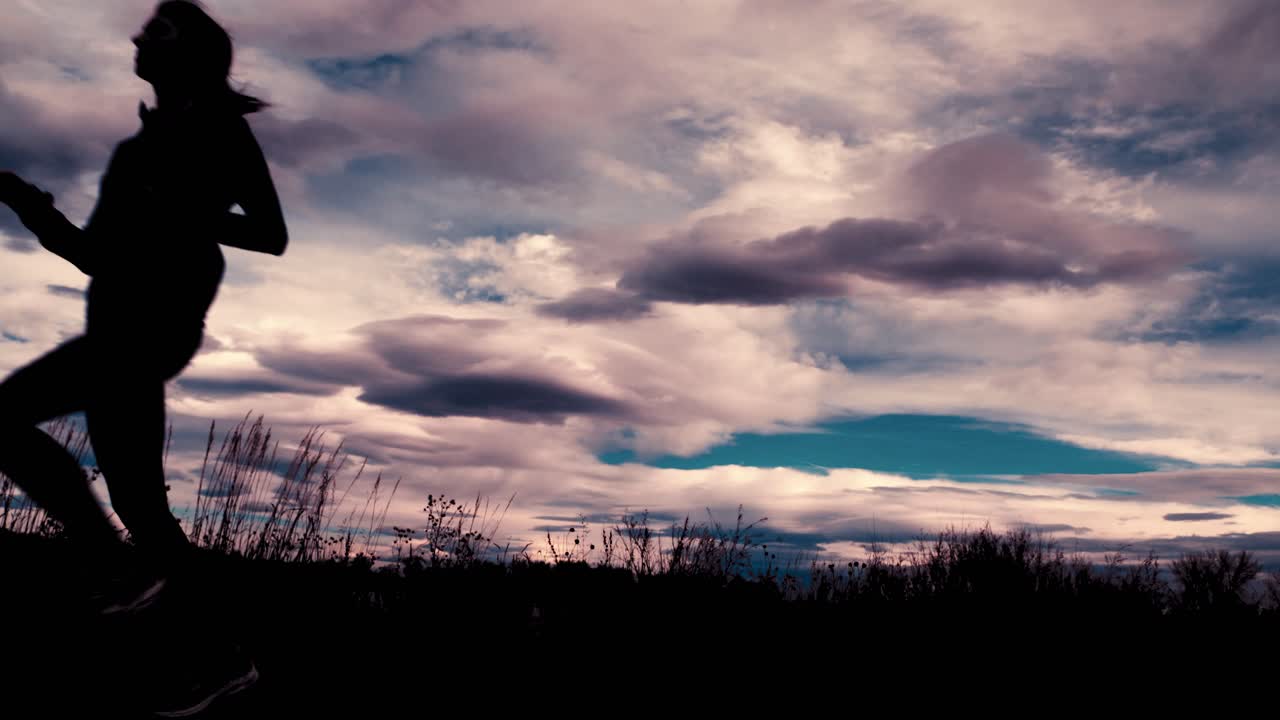 Beautiful black silhouette outline of a female jogger, jogging against a dramatic sky.  The jogger runs toward the camera from right to left.  A wide angle frames this stunning clouds and scenery.
