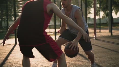 Primer-Plano-De-Un-Hábil-Jugador-De-Baloncesto-Botando-El-Balón-Entre-Las-Piernas,-Mientras-Que-Un-Segundo-Jugador-Defiende-En-Una-Cancha-De-Baloncesto-Al-Aire-Libre-1