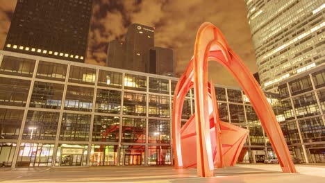 Timelapse-captures-an-imposing-large,-twisted-orange-sculpture-situated-against-a-modern-grid-patterned-building-under-a-dark,-cloudy-sky