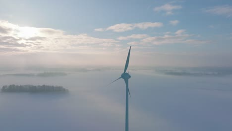 Aerial-view-of-wind-turbines-generating-renewable-energy-in-the-wind-farm,-snow-filled-countryside-landscape-with-fog,-sunny-winter-evening-with-golden-hour-light,-wide-drone-shot-moving-backward