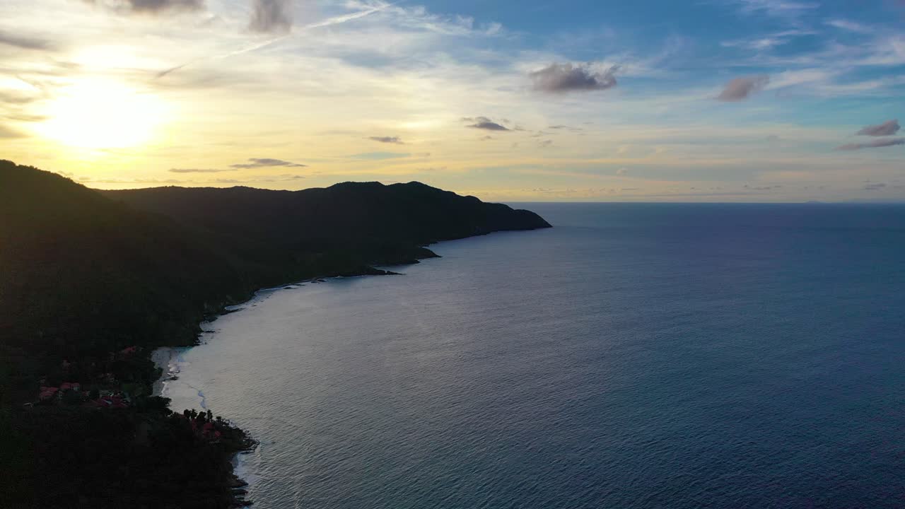 Drone footage captures St. Croix’s calm Caribbean waters at dawn, with a radiant sky of vibrant colors illuminating a tree-covered peninsula and casting soft reflections on the shore