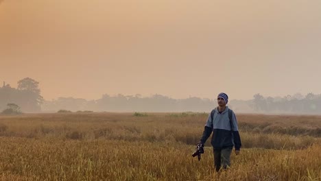 Man-with-DSLR-camera-wonders-through-paddy-field-by-Gas-Plant,-tracking-shot