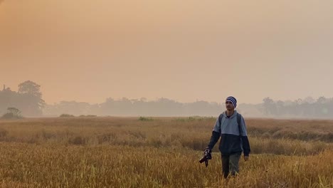 man with dslr camera wonders through paddy field by gas plant, tracking shot