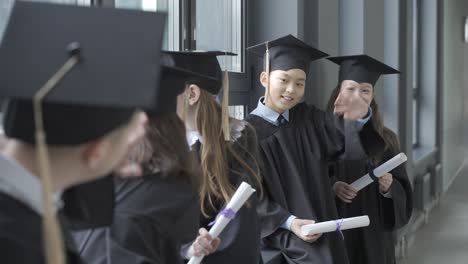 group of happy multiracial preschool students in mortarboard and gown. they are talking and holding diplomas. one of the students says hi to another one.