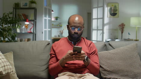 african american man sitting on sofa and typing on phone