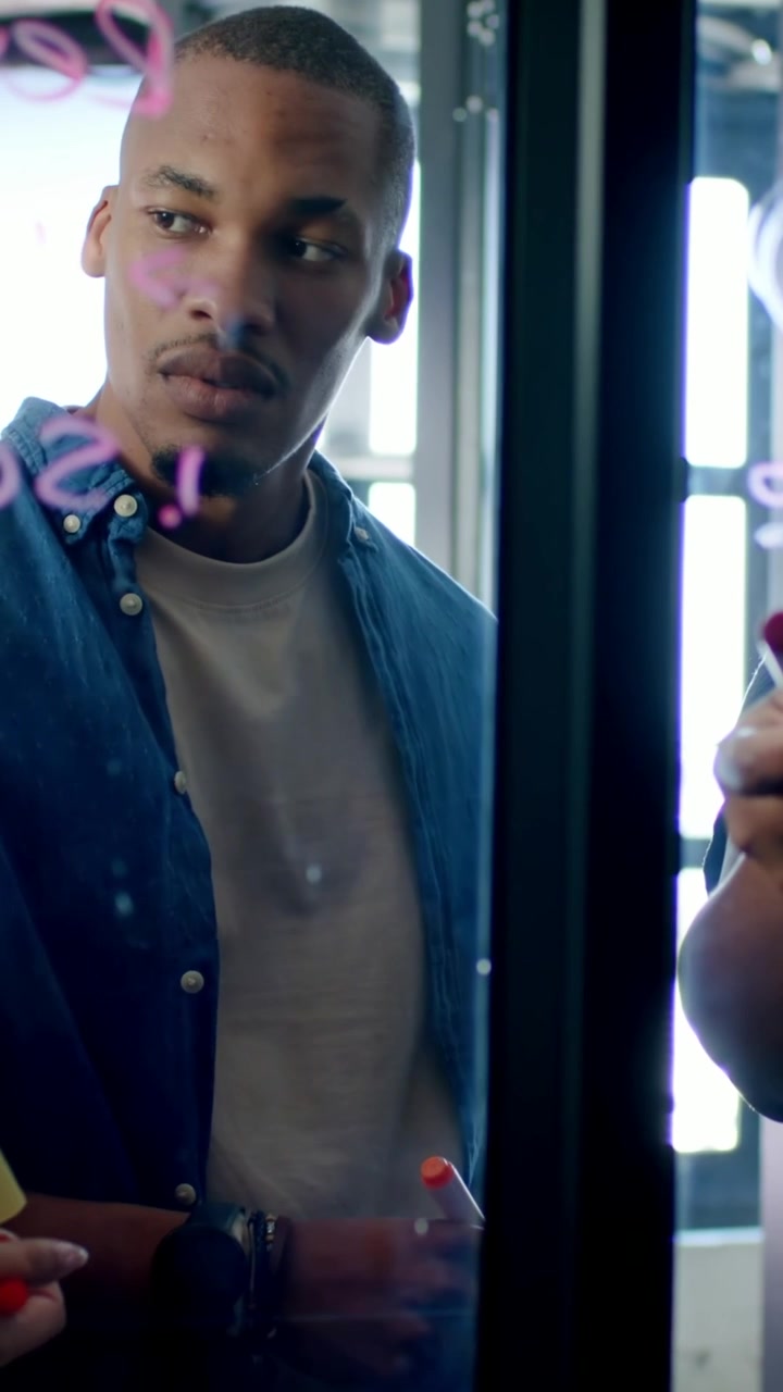 Video verticale: Uomo in denim osserva tenendo un pennarello arancione mentre un collega scrive in rosa sul vetro, pronto.