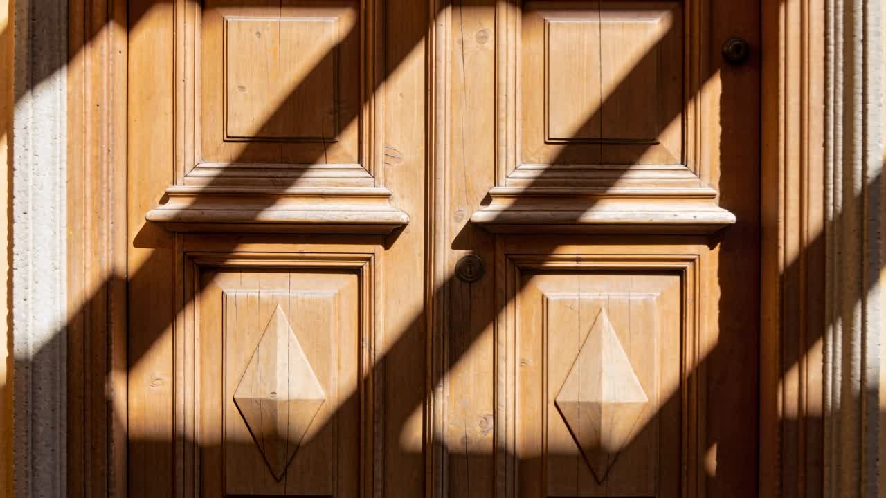 Caméra reculant révélant une double porte en bois sculpté dans un encadrement en pierre, avec une serrure et des ombres glissantes.