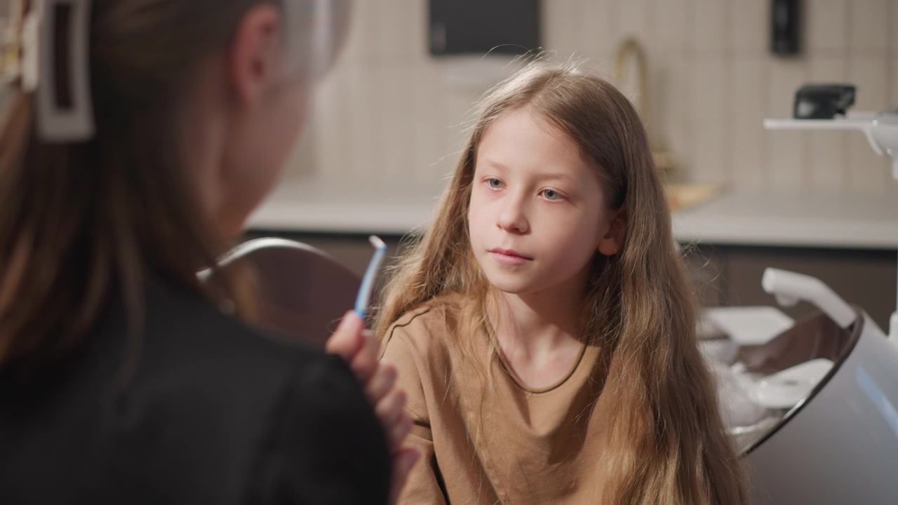 Close up of young girl sitting in orthodontic clinic while interacting with her dentist. scene shows dental professional holding tools and explaining procedures set against soft lighting