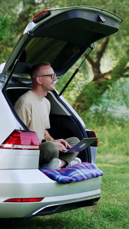 Happy man sits in his car trunk in the forest. Smiling freelancer works on laptop enjoying nature. Vertical video