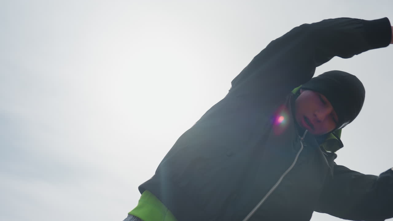 low-angle silhouette caucasian goalkeeper with arms raised against bright sunflare, dramatic backlight and cold sky, hoodie and beanie rimmed by light, triumphant stance on empty urban pitch
