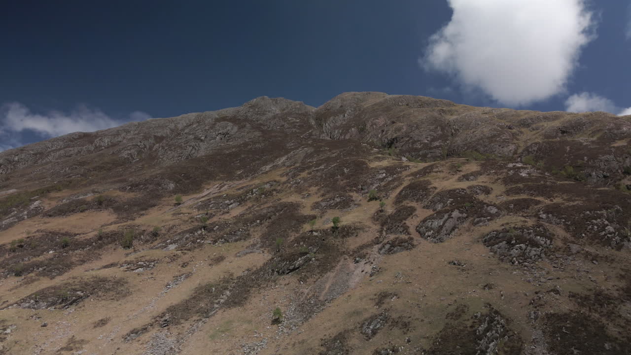 Drone tracking left along side of Scottish mountain looking up, featuring blue sky and dry brown grass