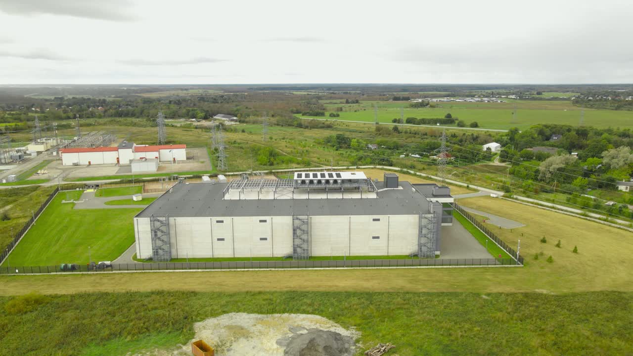 Aerial drone footage orbiting around a large modern data center facility meant for data, AI and server housing in a rural grassy green countryside. Power grid next to it for cybersecurity