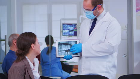 Woman-sitting-on-chair-listening-doctor-looking-on-tablet-with-x-ray