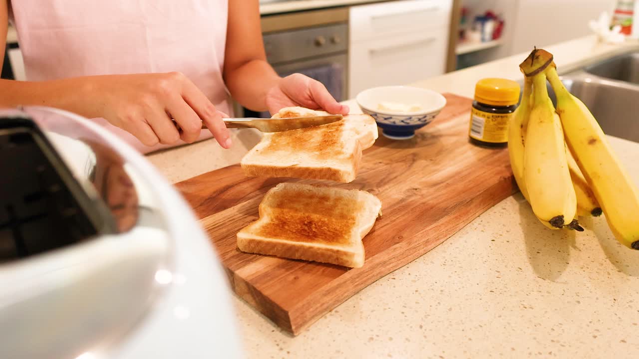 A person spreads butter on toast in a well-lit kitchen, surrounded by bananas and a toaster