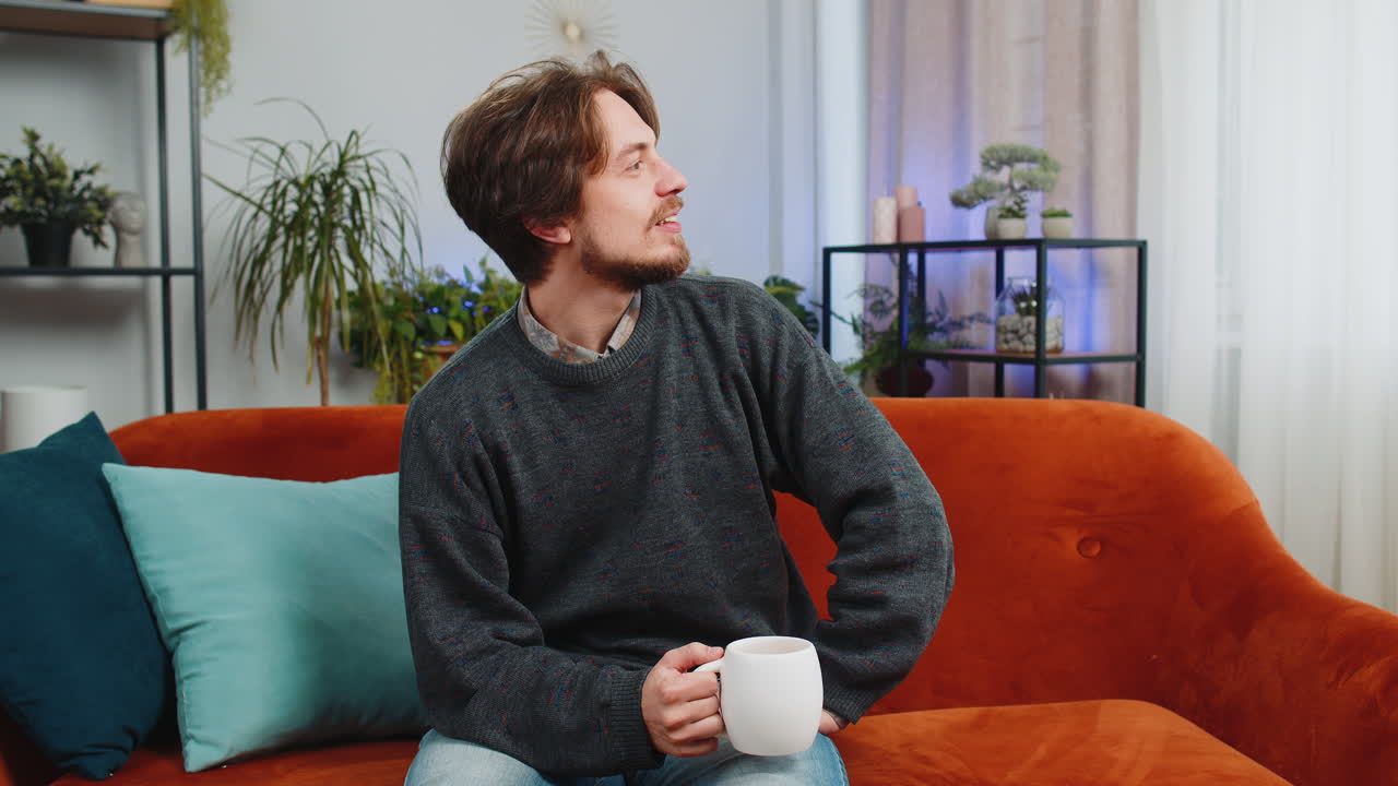 Attractive smiling caucasian young man drinking a cup of coffee or herbal tea sitting at home couch