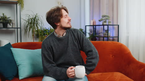 attractive smiling caucasian young man drinking a cup of coffee or herbal tea sitting at home couch