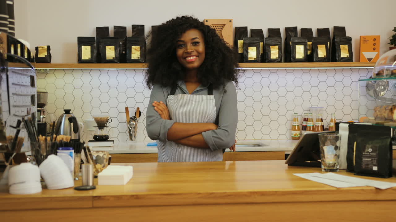 Young african american female barista posing for the camera and crossing arms behind the bar of a coffee shop