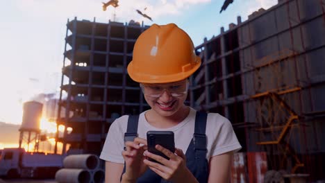 primer plano de una trabajadora asiática con gafas de protección y casco de seguridad sonriendo y usando un teléfono inteligente mientras está de pie en el sitio de construcción