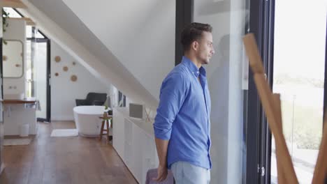 thoughtful biracial man looking out of window in modern apartment, copy space, slow motion