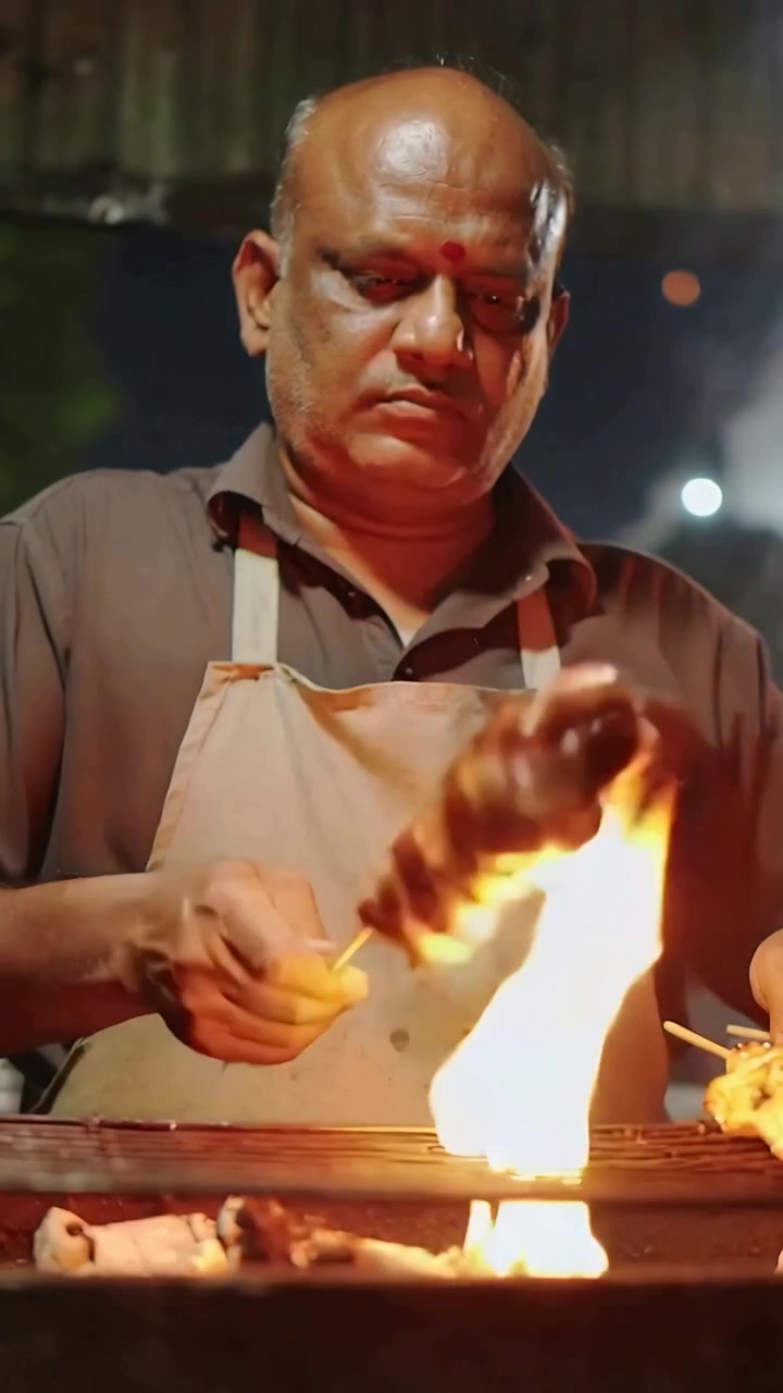 Video verticale: Cuoco che gira e cuoce un kebab su una griglia a carbone di notte, mentre una fiammata di grasso incendia il grembiule.