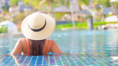 Primer-Plano-De-Una-Mujer-Con-Un-Sombrero-Para-El-Sol-Y-Relajándose-En-La-Piscina-De-Un-Resort-De-Espaldas-A-La-Cámara