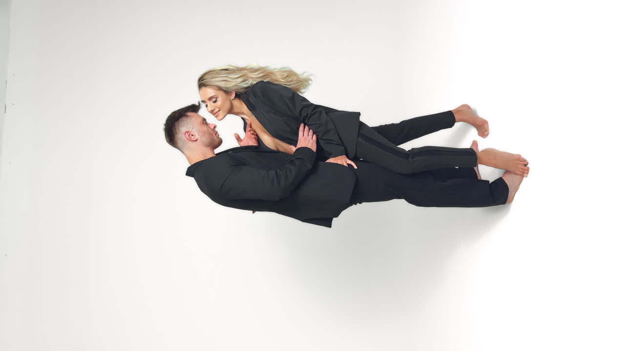 Caucasian young couple wearing black suits stand in studio. Couple stroking each other. White backdrop. Vertical screen.