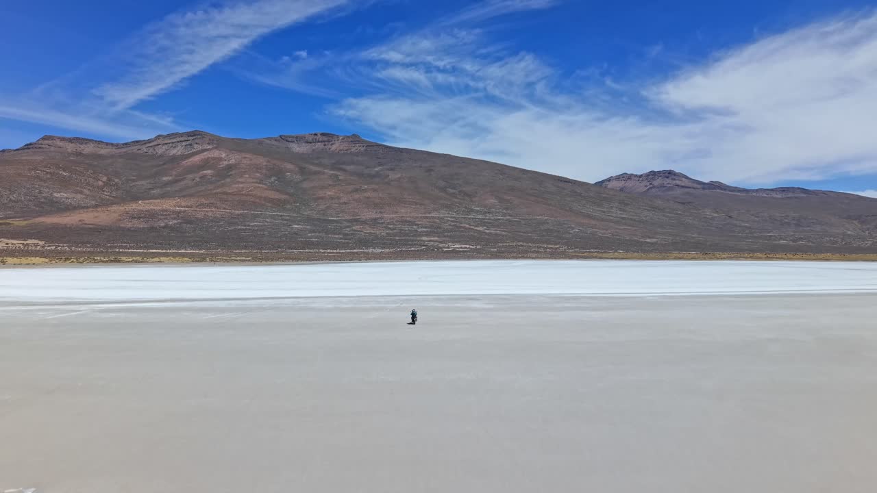 Dois viajantes atravessam as infinitas planícies de sal brancas, com a motocicleta parecendo minúscula na imensa paisagem, cercada por colinas distantes cobertas de sal e céu aberto