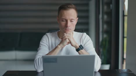 Concentrated young man sits at desk in room and looks attentively at laptop screen