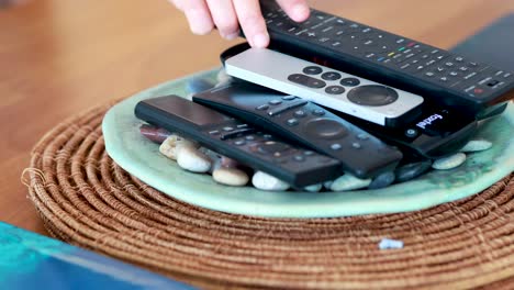 a hand selects and lifts a remote control from a stack arranged on a decorative plate with stones, in a brightly lit modern living room