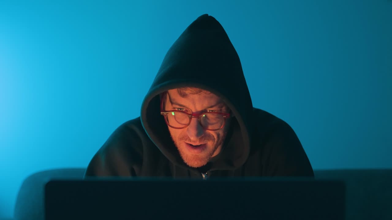 Man in glasses and hoodie sitting behind a laptop in dramatic blue and orange light, cybersecurity, hacking, online fraud, data privacy, secretive computer. online fraud, data privacy, Front view