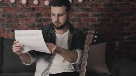 young musician man looking at sheet music to play guitar at home