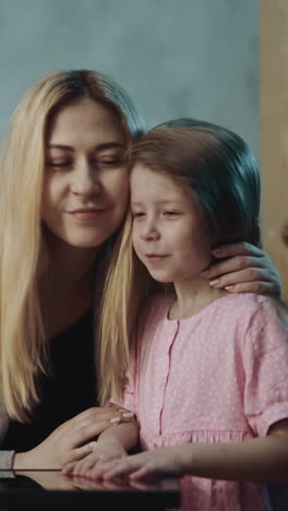 careful long haired woman gives vitamin pill to cute little blonde daughter in pink sitting at small table in semi-dark room at home close view slow motion