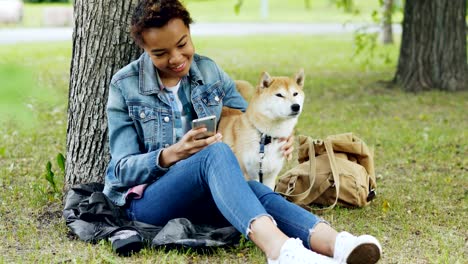 happy african american woman is using smartphone and caressing her cute pet dog resting in city park on windy summer day. nature, animals and people concept.