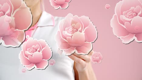 Animation-of-pink-roses-floating-over-hands-of-woman-holding-breast-cancer-ribbon,-on-pink
