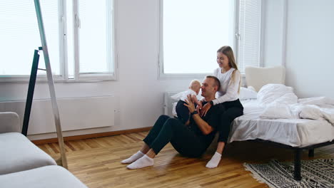 woman sits on bed and her husband sits at her feet. happy dad rises tiny baby above the head.