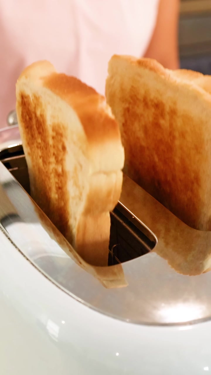 Slices of bread toast in a shiny toaster, popping up as a person stands nearby in a well-lit kitchen