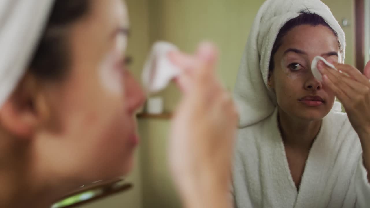 Portrait of biracial woman with vitiligo wearing bathrobe, looking in mirror and cleansing face
