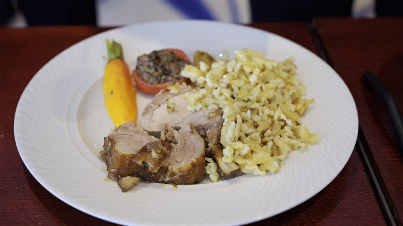 Close up of a server arranging a portion of egg noodle near a few pieces of pork, cooked carrot and stuffed tomato on a white plate
