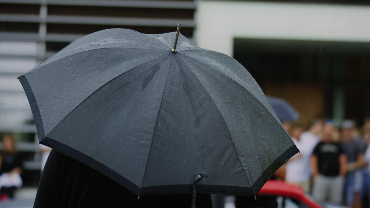 클로즈업 에서, 한 사람 이 검은 우산  ⁇  에 숨어 있는 것 을 볼 수 있으며, 다른 여러 사람 들 이 배경 에 서 있다