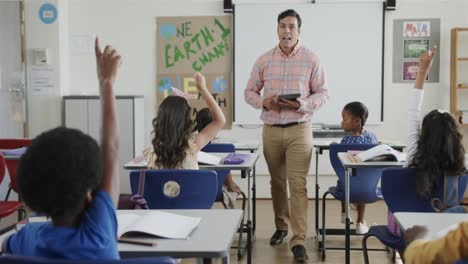 diverse children raising hands and male teacher with tablet in elementary school class, slow motion