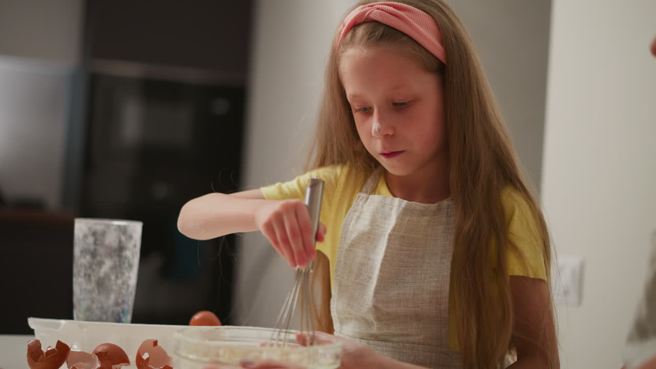 close up of young girl in yellow shirt and apron whisking flour in glass bowl as adult supports bowl from below, broken eggshells scattered on kitchen table and flour dust visible on nose and fingers